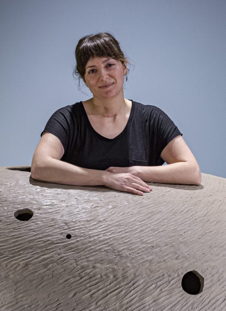 Marisa smiles softly with her arms folded over a huge textured clay sculpture.