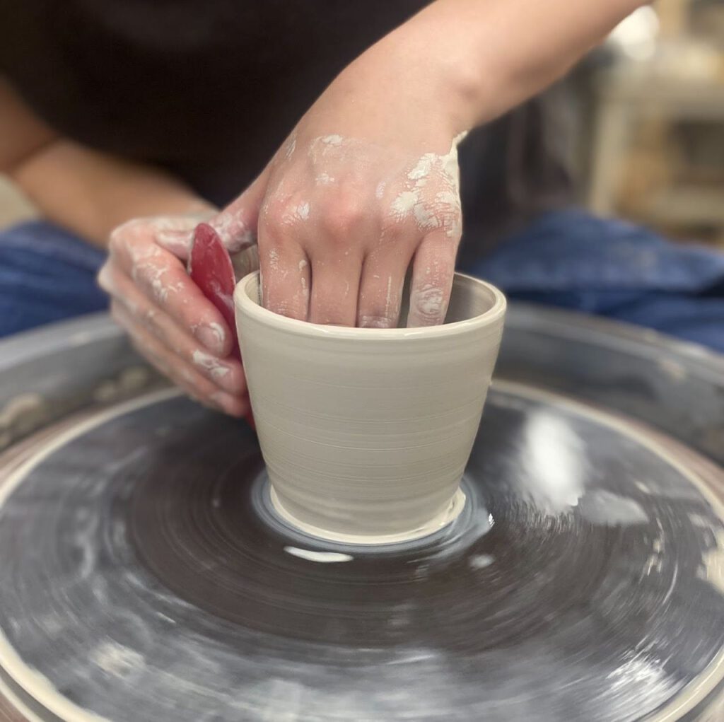 One hand inside a clay vessel with the other flattening the sides with a tool